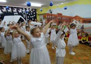 Na tle zimowej dekoracji dzieci przebrane za Anioły tańczą w rzędach z rękoma uniesionymi w górę.